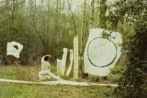 An artist is performing martial arts in the forest. He is surrounded by white paper of different heights and sizes. The largest piece to his right paints a large circle in black ink.